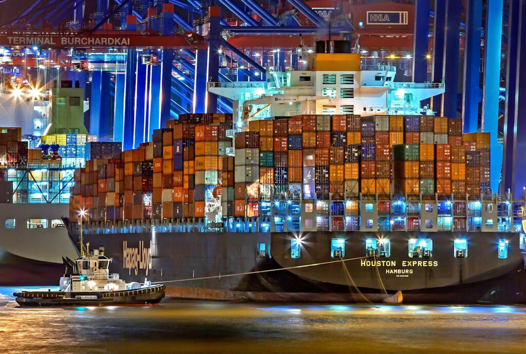 Cargo ship with containers docked at a shipping terminal with cranes in the background.
