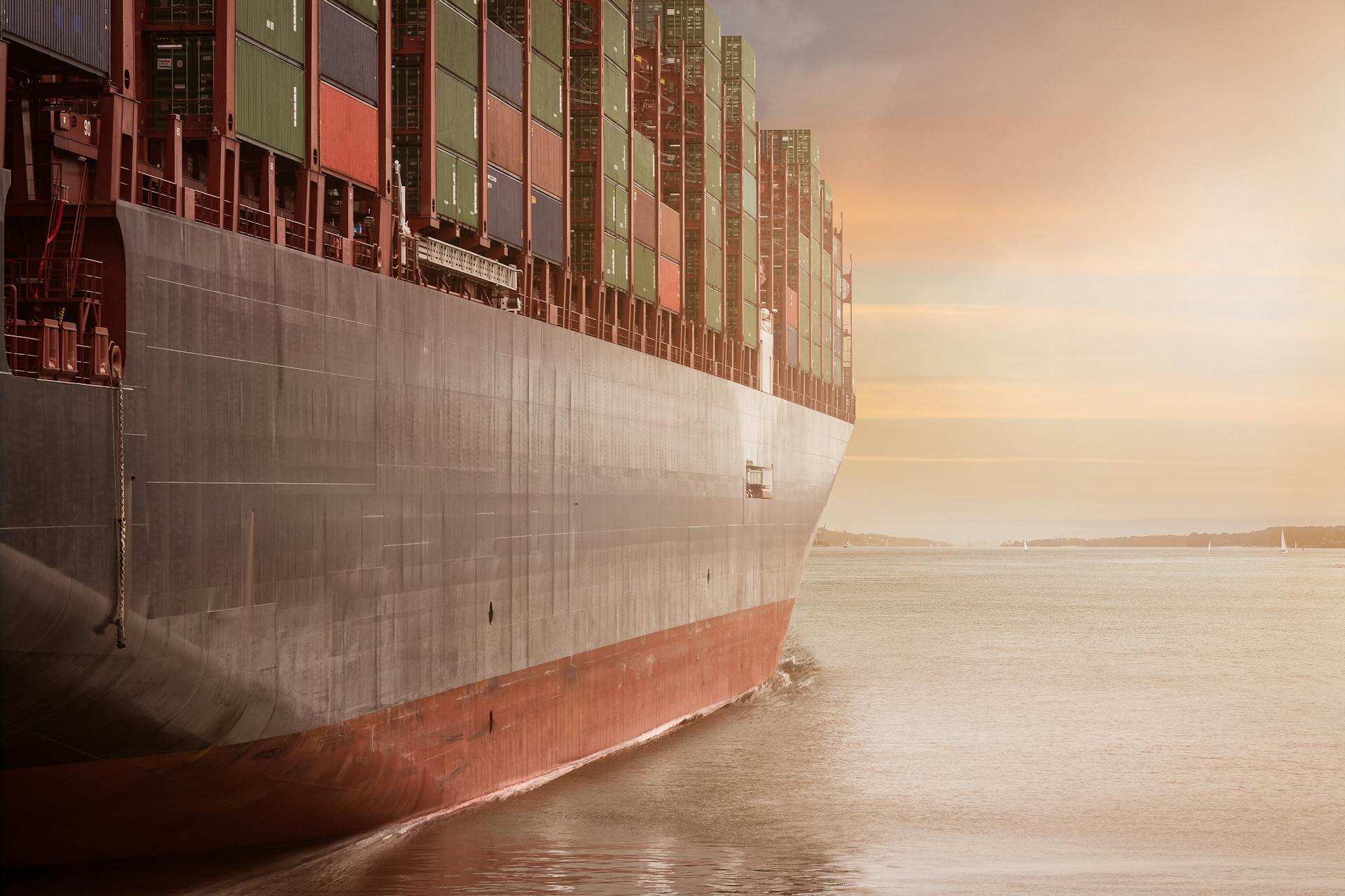 A cargo ship on the water with mountains and cloudy skies in the background.
